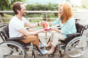 Harmonious relations. Side view on a romantic husband and wife holding hands together and smiling cheerfully while sitting in wheelchairs and celebrating something.