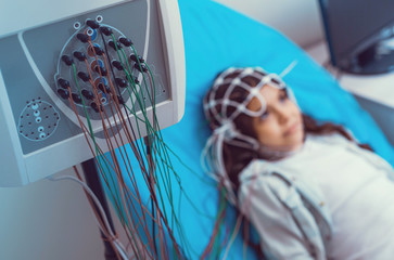 Serious diagnostics. Selective focus on an electroencephalography machine with nodes analyzing brain processes of a child lying on an examination couch in the background.