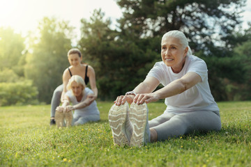 No back pain. Happy retired lady sitting on the grass and reaching for her toes while exercising in a local park.