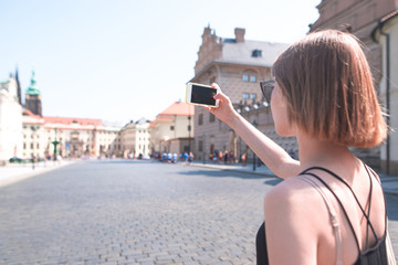 Fototapeta na wymiar Woman in a black dress walks on a sunny summer day along the streets of the old town and makes a photo on a smartphone. Tourist girl photographs the streets of Prague, Czech Republic