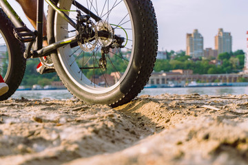 a young guy on a bicycle with big wheels is riding on the river bank in the sand against the backdrop of a big city on a clear hot summer day