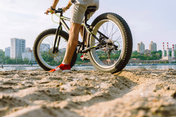 Fototapeta na wymiar a young guy on a bicycle with big wheels is riding on the river bank in the sand against the backdrop of a big city on a clear hot summer day