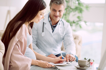 Medical agreement. Joyful positive pregnant woman smiling and signing the document while sitting...