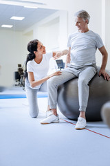 Obraz premium Be attentive. Smiling clever experienced trainer looking at the cheerful ill aged man in a rehabilitation center and showing him the correct way of doing important exercises on a lovely big fitball
