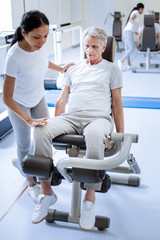 Very careful. Calm serious senior man looking attentive while sitting on a special exercise machine with a qualified reliable specialist of a rehabilitation center examining his injured knee