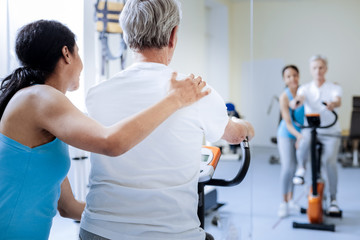 Little help. Kind attentive supporting medical worker putting her hand on a shoulder of a positive enthusiastic aged man while helping him to train on an exercise bike
