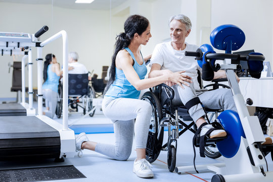 Feeling glad. Cheerful optimistic aged disabled man sitting in wheelchair and looking at the kind attentive qualified specialist before starting the exercises