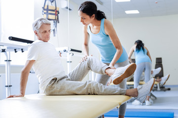 Training muscles. Concentrated medical worker of a progressive modern rehabilitation center bending the injured knee of her tired worried aged patient and looking attentively at him