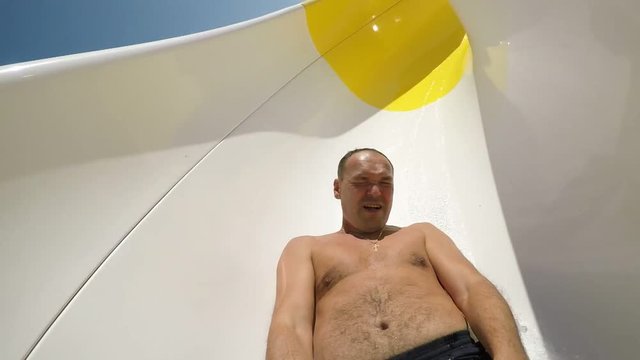 An exciting view of a cheery man sliding dowm on yellow tube in aqua park in summer. He smile and shoots with a selfie stick in slow motion.