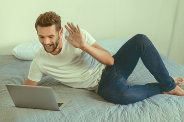 Hello there. Cheerful young man in casual lying on his bed and waving his hand while welcoming his friend during a video call on a laptop.