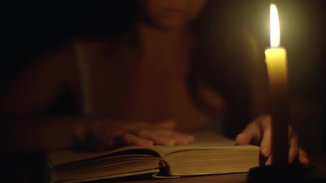 adult woman reading a book in the dark under the light of a burning candle