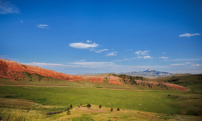 A highway winding through green hills and red rock walls with mountains in the distance along Chief Joseph highway in a sunny afternoon Wyoming landscape