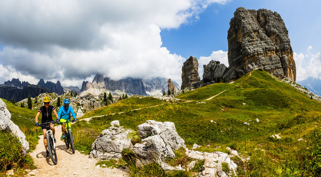 Tourist cycling in Cortina d'Ampezzo, stunning Cinque Torri and Tofana in background. Woman and man riding MTB trail. South Tyrol province of Italy, Dolomites.