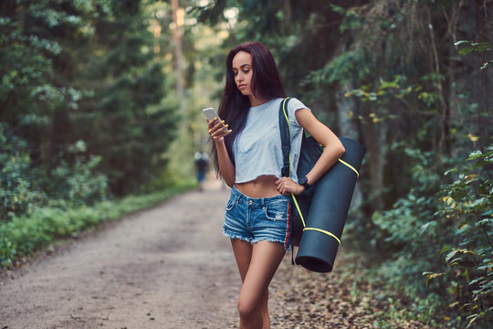 Hipster girl dressed in a shirt and shorts with tourist mat and backpack using a smartphone while standing in a summer forest.