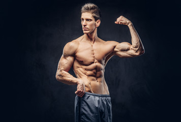 Fototapeta na wymiar Handsome shirtless man with stylish hair and muscular ectomorph shows his bicep on the dark textured background.
