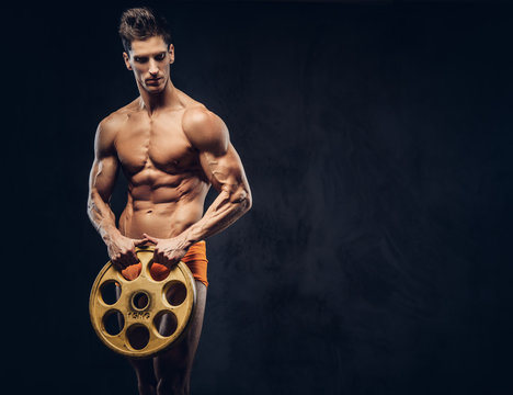 Handsome naked ectomorph bodybuilder with stylish hair in underpants posing with a barbell disk on a dark background.