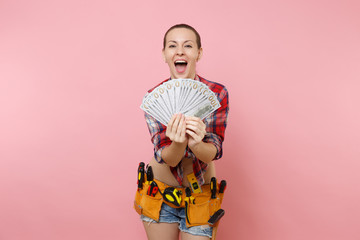 Excited handyman woman in plaid shirt, denim shorts, kit tools belt full of variety instruments hold bundle of cash dollar money isolated on pink background. Female doing male work. Renovation concept
