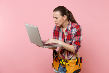 Puzzled stress handyman woman user in plaid shirt, denim shorts, kit tools belt full of instruments working on laptop pc computer isolated on pink background. Female in male work. Renovation concept.