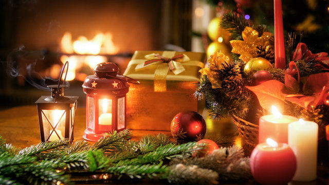 Closeup image of golden gift box, candles and lanterns against Christmas tree and fireplace