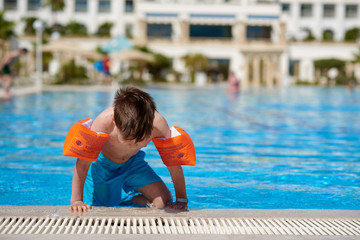 Cute Caucasian boy in floating sleeves getting out from swimming pool at resort.