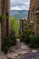 Fototapeta na wymiar A view of a narrow, stone slab road through the walls of very old buildings, a mountain and populated area in the distance, there are many dark clouds in the sky