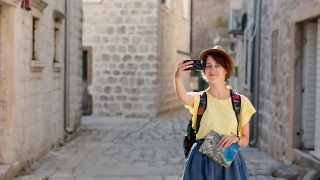 Cheerful female tourist in hat, make a picture on modern cell phone device walking on streets. Positive hipster traveller walking in European city. Sunny day in Montenegro.