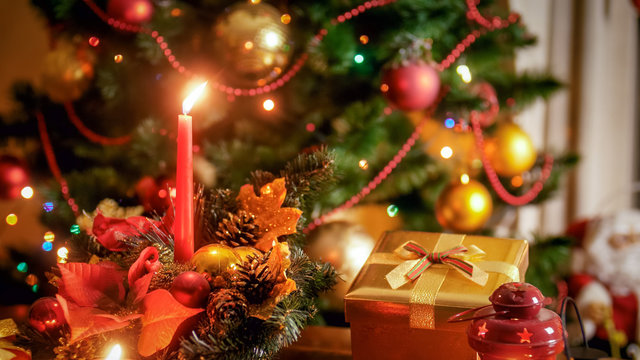 Closeup image of golden gift box and burning candle against beautiful Christmas tree