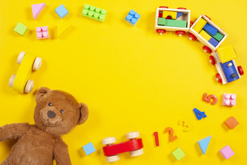 Baby kids toys background with teddy bear, wooden train, colorful blocks and bricks on yellow background. Top view