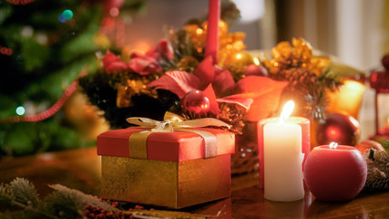 Fototapeta na wymiar Closeup image of gift box with golden ribbon and burning candles against Christmas tree at living room