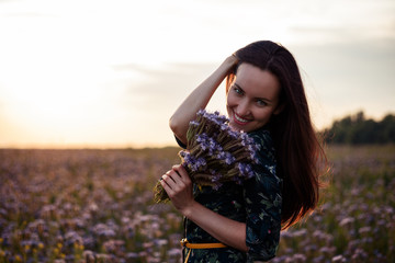 Portrait of a young woman in a flower meadow at sunset with a bouquet of purple flowers, smiling, copy space