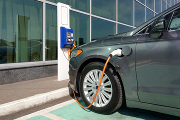 Charging electic car with plugged supply cable at electro vehicle charge station. Eco power machine parked at modern office building with glass window facade