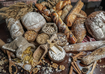 Bread background. Brown and white whole grain loaves wrapped in kraft paper composition on rustic dark wood with wheat ears scattered around. Baking and home bread making concept. Soft toning
