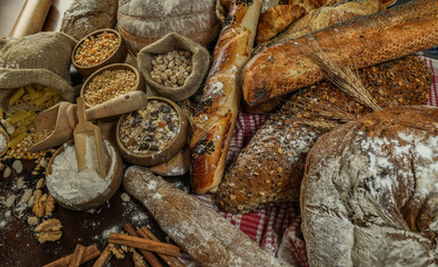 Bread background. Brown and white whole grain loaves wrapped in kraft paper composition on rustic dark wood with wheat ears scattered around. Baking and home bread making concept. Soft toning
