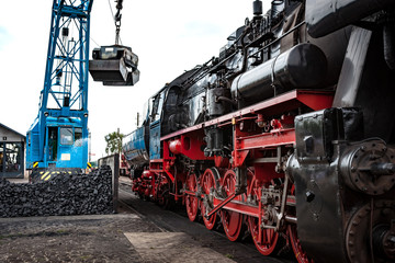 Obraz premium Steaming steam train or locomotive is waiting for coal loading by a vintage crane, industrial details and craftsmanship along the rail way and train station