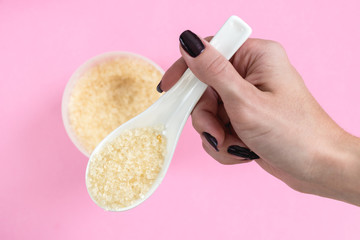 Close up on a female hand holding a white ceramic spoon with bath salt and a blurred jar of bath salt in the background.