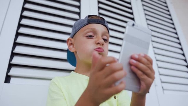 Portrait of a funny little boy looks in the phone, shows a fist, inflates his lips, gets angry. Internet network, online communication, social network. Child and modern technology
