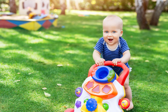 Cute Little Boy Learning To Walk With Walker Toy On Green Grass Lawn At Backyard. Baby Laughing And Having Fun Making First Step At Park On Bright Sunny Day Outdoors. Happy Childhood Concept