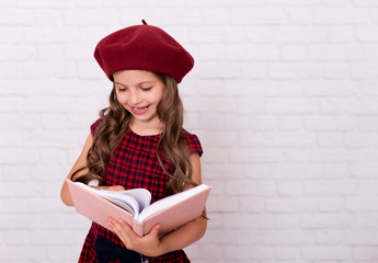  Portrait of a smiling little schoolgirl  in a red beret holding open pink notebook.