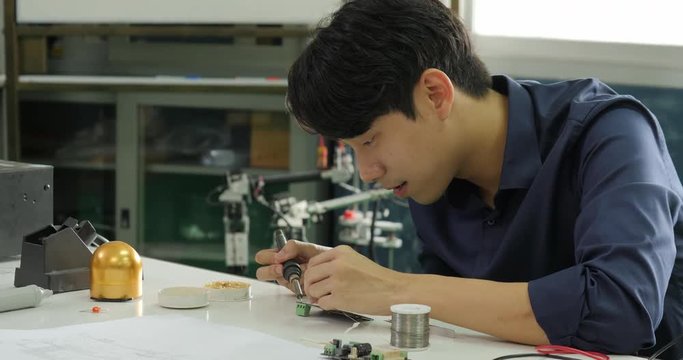 Asian electronics engineer is soldering an electric board in his workshop. Asian student are studying electronics and soldering in classroom.