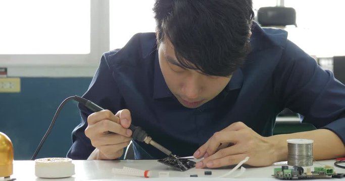 Asian electronics engineer is soldering an electric board in his workshop. Asian student are studying electronics and soldering in classroom.