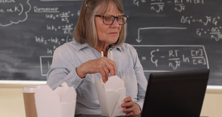 Elderly female teacher having lunch and using laptop in classroom setting