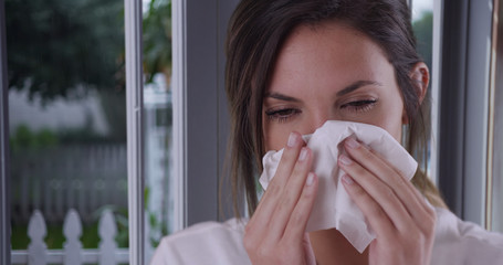 Female with cold or allergies wiping nose with tissue inside house by window