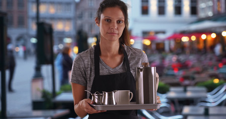 Unhappy waitress wearing apron and carrying tray with coffee at outdoor cafe