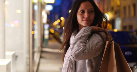 Beautiful female carrying shopping bags looking around on city street at night