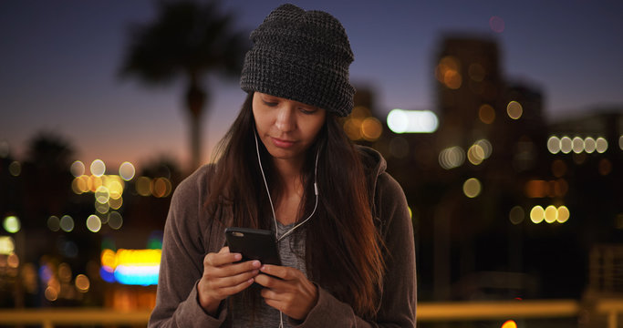 Millennial Girl In The City At Night Texting And Listening To Music On Cellphone