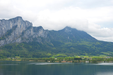A mountain dwarfs a small village which is on the edge of a lake. Storm clouds fill the sky. The lower slopes of the mountain are covered in forest.