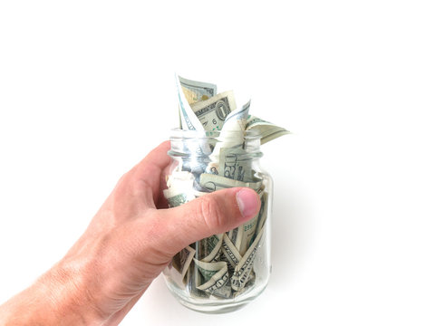 A closeup photograph of a white Caucasian male hand holding a glass mason savings jar with wads of cash hanging out of the top isolated on a white background.