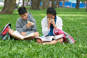 Teenage school children eating sandwiches and reading books outdoors