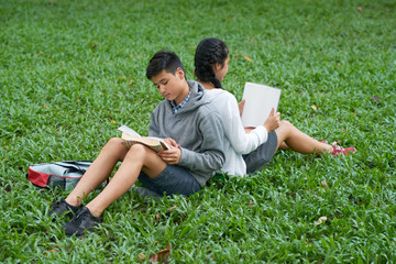 Naklejka na ściany i meble Asian school children sitting in park back to back and reading books