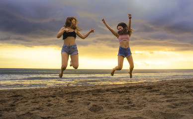 lifestyle portrait of two happy and attractive young Asian Korean women girlfriends jumping on sunset beach excited and cheerful enjoying summer holidays